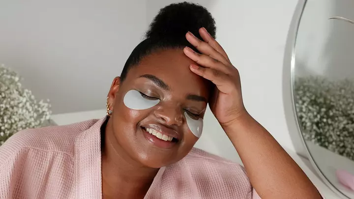 A curvy African-American woman in a pink robe relaxing with hydrating under eye patches